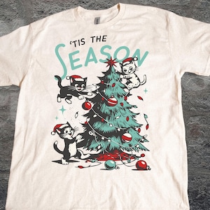 Può includere: T-shirt color crema con un motivo natalizio. Presenta un albero di Natale decorato con palline rosse e luci, con tre gatti neri con cappelli da Babbo Natale. Il testo "'Tis the Season" è sopra l'albero.