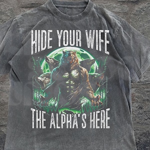 May include: Dark gray t-shirt featuring a werewolf design with the text "HIDE YOUR WIFE THE ALPHA'S HERE". The graphic includes a full moon and forest scene. The shirt has a vintage, distressed look.