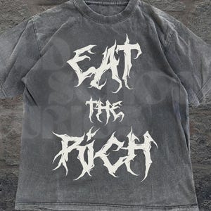 May include: A gray t-shirt with the text "EAT THE RICH" in a white, gothic font. The shirt has a vintage, washed appearance. The words are stacked vertically on the front of the shirt. The brand "OLD SCHOOL CURIOS" is printed at the bottom right.