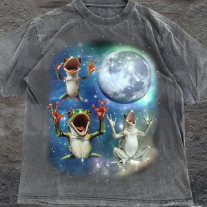 May include: Gray t-shirt featuring a whimsical design of three frogs with open mouths and raised arms, set against a starry night sky and a large moon. The shirt has a vintage, washed look, with the text "OLD SCHOOL CURIOS" at the bottom.