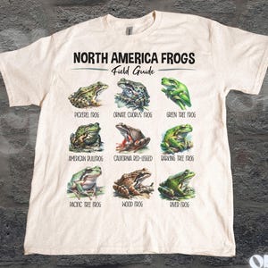 May include: Cream-colored t-shirt featuring a field guide of North American frogs. The shirt displays nine different frog species with their names, including Pickerel Frog, Green Tree Frog, and Pacific Tree Frog. The text "North America Frogs Field Guide" is printed at the top.
