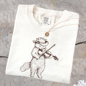 May include: Cream-colored t-shirt featuring a detailed illustration of a cat wearing a cowboy hat and playing a violin. A gold-colored necklace with a pendant rests on the shirt. The shirt has the Comfort Colors brand label.