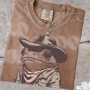 May include: Brown Comfort Colors t-shirt featuring a cartoon frog wearing a cowboy hat, jacket, and neckerchief. A silver necklace with a small pendant rests on the shirt. The shirt has the Comfort Colors tag and the text "Old School Curios".