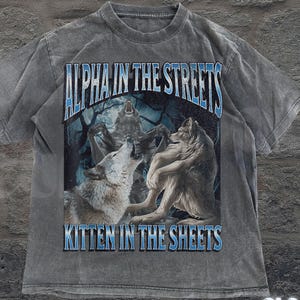 May include: Gray t-shirt with a wolf graphic. The shirt features the text "ALPHA IN THE STREETS" above a wolf illustration and "KITTEN IN THE SHEETS" below. The shirt also has the text "OLD SCHOOL CURIOS" at the bottom.