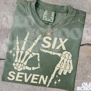 May include: An olive green t-shirt with a skeleton hand graphic displaying the numbers "SIX" and "SEVEN". A silver necklace with a small pendant rests on the shirt. The shirt is folded on a textured surface.