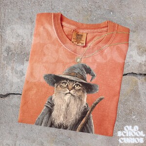 K&ouml;nnte beinhalten: Orangefarbenes Comfort Colors T-Shirt mit einer Katze als Zauberer, langem Bart und Stab. Eine goldene Halskette mit Anh&auml;nger liegt auf dem Shirt. Das Shirt hat ein Etikett mit der Aufschrift "Comfort Colors". Der Text "Old School Curios" befindet sich unten rechts.