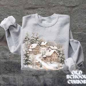 Op de afbeelding: Een lichtgrijs sweatshirt met een winterdorpssc&egrave;ne met besneeuwde huizen en groenblijvende bomen. Het ontwerp is in aquarel stijl. De tekst "OLD SCHOOL CURIOS" staat rechtsonder.