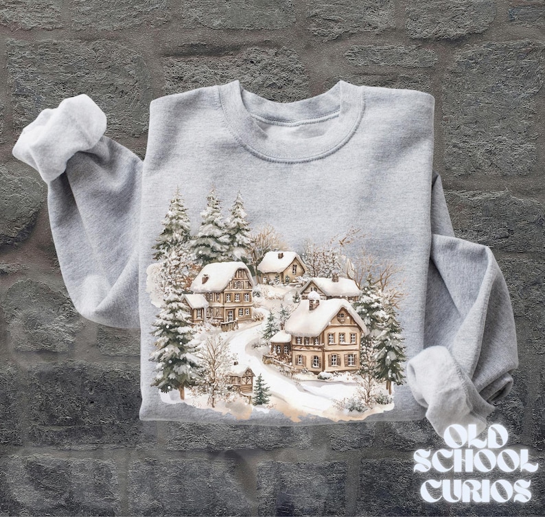 Op de afbeelding: Een lichtgrijs sweatshirt met een winterdorpssc&egrave;ne met besneeuwde huizen en groenblijvende bomen. Het ontwerp is in aquarel stijl. De tekst "OLD SCHOOL CURIOS" staat rechtsonder.