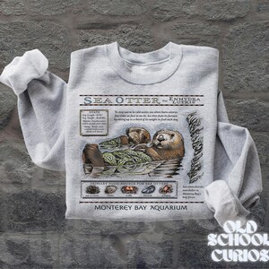 May include: A light grey sweatshirt with a detailed illustration of sea otters, scientific information, and the text "SEA OTTER - ENHYDRA LUTRIS." The sweatshirt also includes the text "Monterey Bay Aquarium" and "Old School Curios."