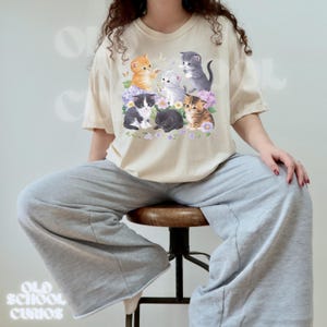 以下が含まれることがあります： クリーム色のTシャツ。様々な色の7匹の子猫のグラフィックが描かれ、紫色の花と蝶に囲まれています。「OLD SCHOOL CURIOS」の文字がヴィンテージスタイルでプリントされています。グレーのスウェットパンツと合わせています。