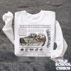 May include: White sweatshirt with a sea otter illustration and the text "SEA OTTER ~ ENHYDRA LUTRIS". The design includes facts about sea otters and their diet. The sweatshirt is from the Monterey Bay Aquarium.