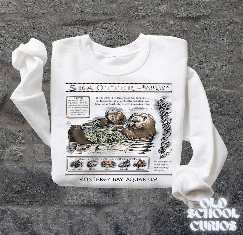 May include: White sweatshirt with a sea otter illustration and the text "SEA OTTER ~ ENHYDRA LUTRIS". The design includes facts about sea otters and their diet. The sweatshirt is from the Monterey Bay Aquarium.
