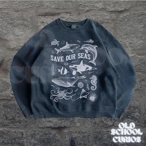 Puede incluir: Sudadera azul oscuro con un gr&aacute;fico blanco de varias criaturas marinas, incluyendo una ballena, un tibur&oacute;n y un delf&iacute;n. El texto "SAVE OUR SEAS" se muestra prominentemente. La sudadera tiene un aspecto vintage.