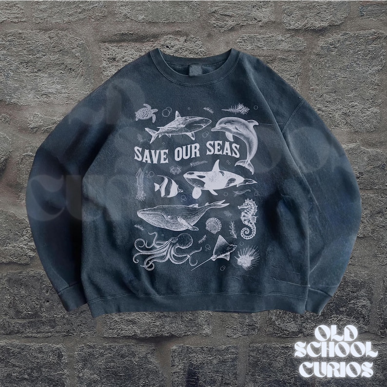 Puede incluir: Sudadera azul oscuro con un gr&aacute;fico blanco de varias criaturas marinas, incluyendo una ballena, un tibur&oacute;n y un delf&iacute;n. El texto "SAVE OUR SEAS" se muestra prominentemente. La sudadera tiene un aspecto vintage.