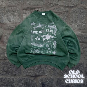 Puede incluir: Sudadera verde con el texto "SAVE OUR SEAS" e ilustraciones blancas de criaturas marinas, incluyendo una ballena, un tibur&oacute;n y un caballito de mar. La sudadera tiene cuello redondo y mangas largas.