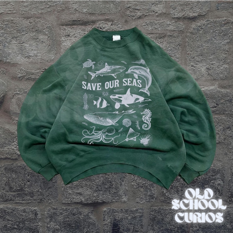Puede incluir: Sudadera verde con el texto "SAVE OUR SEAS" e ilustraciones blancas de criaturas marinas, incluyendo una ballena, un tibur&oacute;n y un caballito de mar. La sudadera tiene cuello redondo y mangas largas.
