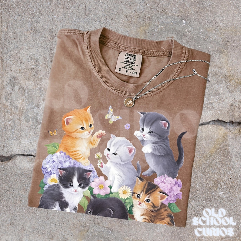 以下が含まれることがあります： 6匹の子猫、花、蝶のグラフィックが特徴の茶色のTシャツ。 シャツはクルーネックと半袖です。 ペンダント付きのシルバーネックレスがシャツの上にドレープされています。 タグには「Comfort Colors」と書かれています。