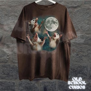 May include: Brown t-shirt featuring a graphic of three opossums howling at a full moon. The opossums are white and gray, with their mouths open. The moon is large and detailed. The shirt has a vintage, distressed look.
