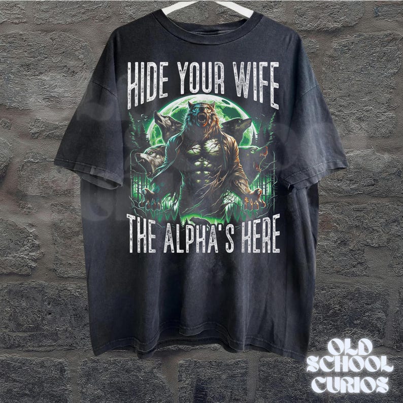May include: Black t-shirt featuring a werewolf illustration with the text "HIDE YOUR WIFE THE ALPHA'S HERE". The graphic includes a werewolf, wolves, and a full moon. The shirt is hanging on a white hanger.