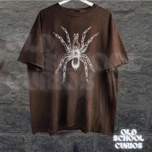 Peut inclure: T-shirt marron avec un motif de tarentule blanche. Le t-shirt est suspendu &agrave; un cintre blanc devant un mur gris textur&eacute;. Les mots "OLD SCHOOL CURIOS" sont imprim&eacute;s en blanc en bas &agrave; droite.