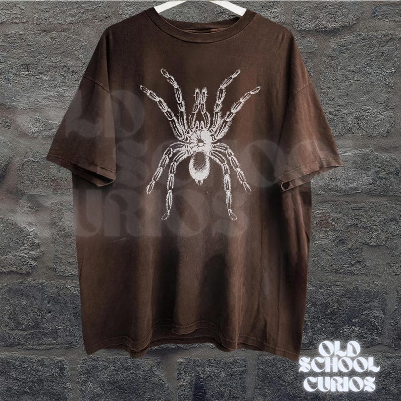 Peut inclure: T-shirt marron avec un motif de tarentule blanche. Le t-shirt est suspendu &agrave; un cintre blanc devant un mur gris textur&eacute;. Les mots "OLD SCHOOL CURIOS" sont imprim&eacute;s en blanc en bas &agrave; droite.