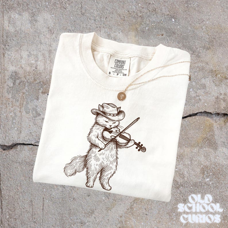 May include: Cream-colored t-shirt featuring a detailed illustration of a cat wearing a cowboy hat and playing a violin. A gold-colored necklace with a pendant rests on the shirt. The shirt has the Comfort Colors brand label.