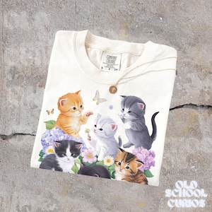 以下が含まれることがあります： クリーム色のTシャツ。6匹の子猫が花と蝶の中にいるグラフィックが描かれています。子猫はオレンジ、グレー、黒など様々な色をしています。金のネックレスとペンダントがシャツの上にあります。シャツにはComfort Colorsのタグが付いています。