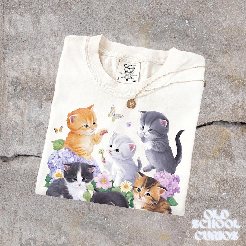 以下が含まれることがあります： クリーム色のTシャツ。6匹の子猫が花と蝶の中にいるグラフィックが描かれています。子猫はオレンジ、グレー、黒など様々な色をしています。金のネックレスとペンダントがシャツの上にあります。シャツにはComfort Colorsのタグが付いています。