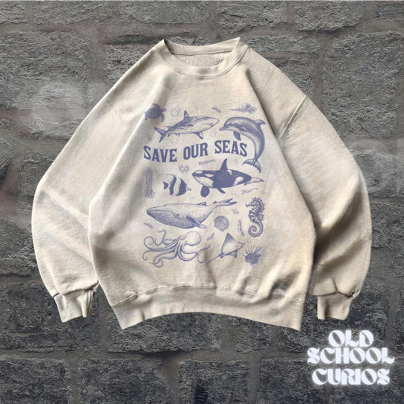 Puede incluir: Sudadera beige con un dise&ntilde;o azul de vida marina, incluyendo una ballena, un delf&iacute;n y un tibur&oacute;n. El texto "SAVE OUR SEAS" est&aacute; impreso sobre las ilustraciones. La sudadera tiene cuello redondo y mangas largas.