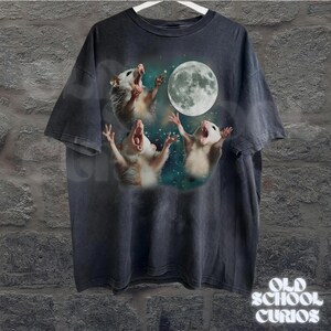 May include: Dark gray t-shirt featuring a graphic of three opossums howling at a full moon. The opossums are white and tan, set against a starry background. The shirt has a vintage, distressed look, with the text "OLD SCHOOL CURIOS" at the bottom.