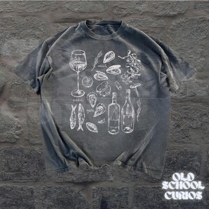 May include: Gray t-shirt with a white illustration of wine, oysters, mussels, sardines, and wine bottles. The text "Old School Curios" is at the bottom right corner of the shirt.