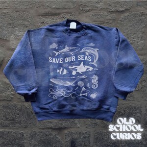 Puede incluir: Sudadera azul con un gr&aacute;fico blanco de varias criaturas marinas, incluyendo una ballena, un tibur&oacute;n y un delf&iacute;n. El texto "SAVE OUR SEAS" est&aacute; impreso encima de las criaturas marinas. La sudadera tiene cuello redondo y mangas largas.