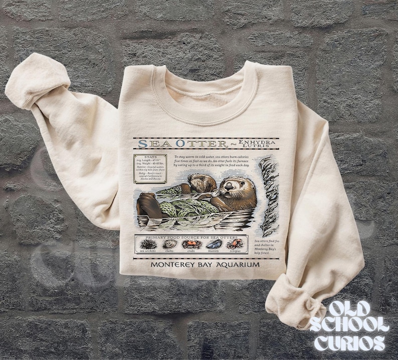 May include: A cream-coloured sweatshirt with a detailed illustration of a sea otter, including text that reads "SEA OTTER ~ ENHYDRA LUTRIS" and "Monterey Bay Aquarium". The graphic includes facts about sea otters and their diet. The sweatshirt has long sleeves.
