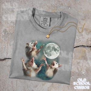 K&ouml;nnte beinhalten: Ein graues Comfort Colors T-Shirt mit einer Grafik von drei Opossums, die den Vollmond anheulen. Eine goldfarbene Sternanh&auml;nger-Halskette liegt auf dem Shirt. Das Shirt hat ein Etikett mit der Aufschrift XL-TG-EG.