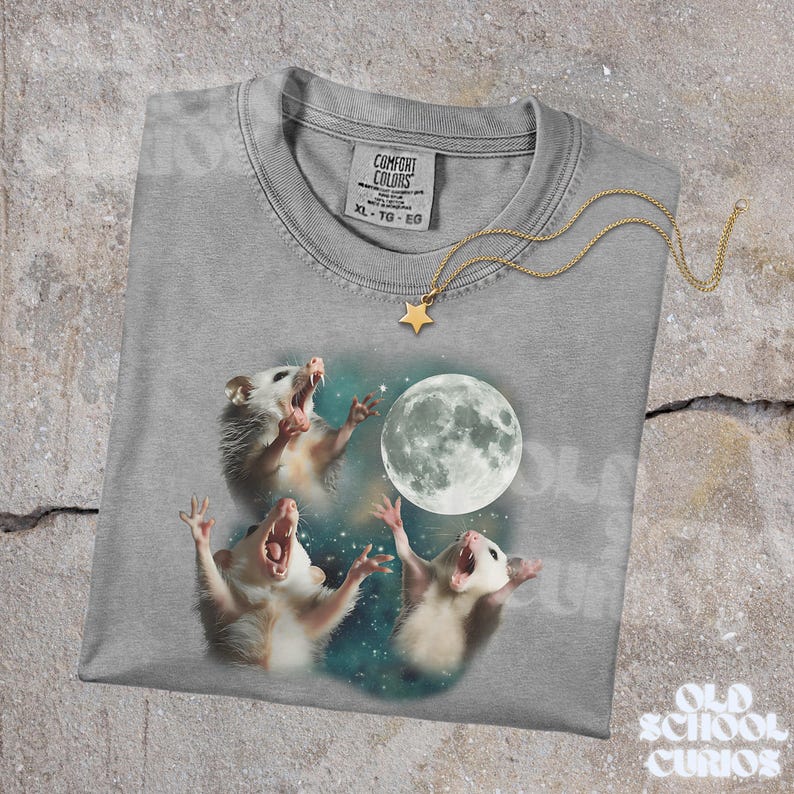 K&ouml;nnte beinhalten: Ein graues Comfort Colors T-Shirt mit einer Grafik von drei Opossums, die den Vollmond anheulen. Eine goldfarbene Sternanh&auml;nger-Halskette liegt auf dem Shirt. Das Shirt hat ein Etikett mit der Aufschrift XL-TG-EG.