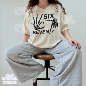 May include: Cream-colored t-shirt with a black skeleton hand graphic forming the number six and the word "SIX" above it. The word "SEVEN" is below the hand graphic, with a black skeleton hand forming the number seven. The shirt is paired with gray sweatpants.