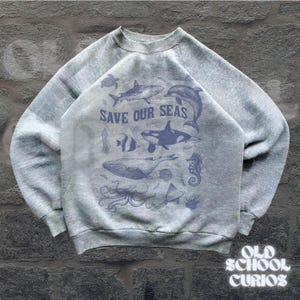 Puede incluir: Sudadera gris con cuello redondo y mangas largas. La parte delantera presenta un gr&aacute;fico azul de varias criaturas marinas, incluyendo una ballena, un tibur&oacute;n y un delf&iacute;n, con el texto "SAVE OUR SEAS". La esquina inferior derecha tiene el texto "OLD SCHOOL CURIOS".