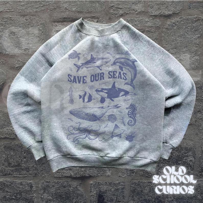 Puede incluir: Sudadera gris con cuello redondo y mangas largas. La parte delantera presenta un gr&aacute;fico azul de varias criaturas marinas, incluyendo una ballena, un tibur&oacute;n y un delf&iacute;n, con el texto "SAVE OUR SEAS". La esquina inferior derecha tiene el texto "OLD SCHOOL CURIOS".