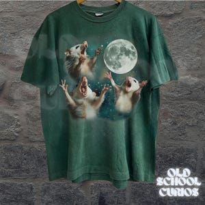 May include: Green t-shirt featuring a graphic of three opossums howling at a full moon against a starry background. The shirt has a vintage, faded look. The text "OLD SCHOOL CURIOS" is printed in white on the lower right.