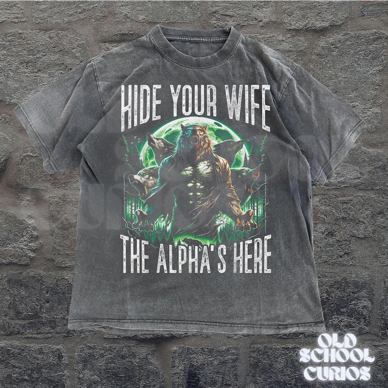 May include: Dark gray t-shirt featuring a werewolf design with the text "HIDE YOUR WIFE THE ALPHA'S HERE". The graphic includes a full moon and forest scene. The shirt has a vintage, distressed look.