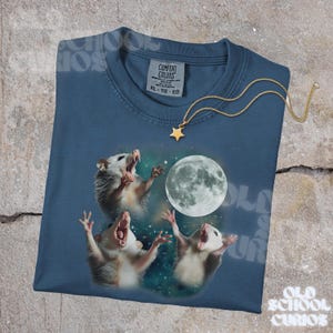 K&ouml;nnte beinhalten: Ein marineblaues T-Shirt mit einer Grafik von drei Opossums, die den Vollmond anheulen. Das Shirt hat einen Rundhalsausschnitt und kurze &Auml;rmel. Eine goldfarbene Sternanh&auml;nger-Halskette ist &uuml;ber dem Shirt drapiert. Der Text "Old School Curios" ist sichtbar.