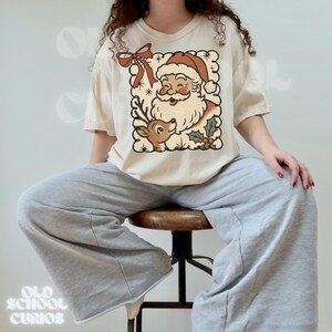May include: Cream-colored t-shirt featuring a vintage-style Santa Claus and reindeer graphic. The design includes a red bow, holly, and snowflakes. The text "OLD SCHOOL CURIOS" is printed at the bottom of the image.