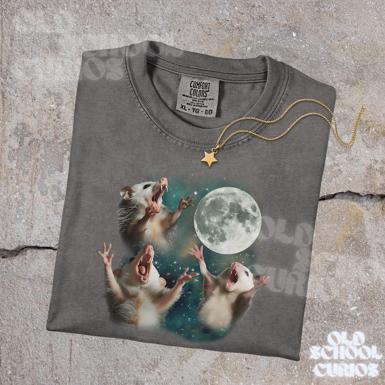 K&ouml;nnte beinhalten: Ein graues Comfort Colors T-Shirt mit einer Grafik von drei Opossums, die den Vollmond anheulen. Eine goldene Sternanh&auml;nger-Halskette liegt auf dem Shirt. Auf dem Shirt steht der Text "OLD SCHOOL CURIOS".