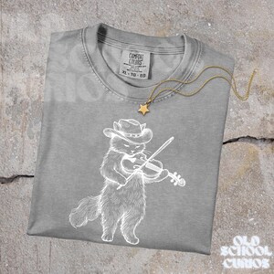 May include: A gray Comfort Colors t-shirt featuring a white illustration of a cat wearing a cowboy hat and playing a violin. A gold star pendant necklace rests on the shirt. The shirt has a tag that reads XL-TG-EG.