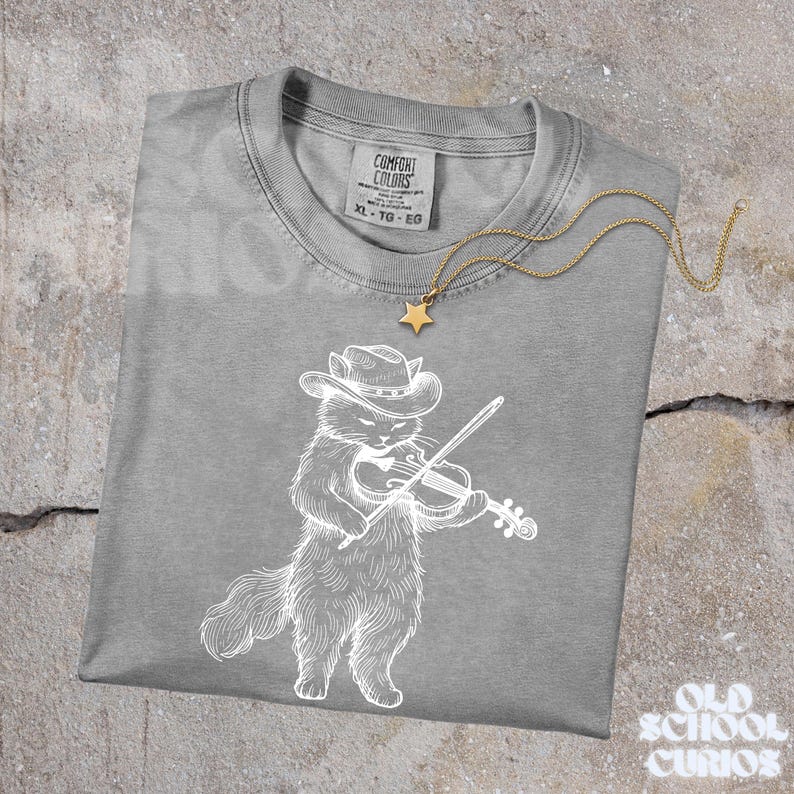 May include: A gray Comfort Colors t-shirt featuring a white illustration of a cat wearing a cowboy hat and playing a violin. A gold star pendant necklace rests on the shirt. The shirt has a tag that reads XL-TG-EG.