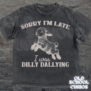 May include: Dark gray t-shirt with the text "SORRY I'M LATE I WAS DILLY DALLYING" and a graphic of a jumping lamb. The shirt has a vintage, distressed look. The brand "OLD SCHOOL CURIOS" is printed at the bottom right.