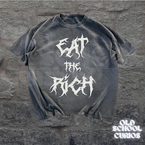 May include: A gray t-shirt with the text "EAT THE RICH" in a white, gothic font. The shirt has a vintage, washed-out look. The words are stacked vertically. The bottom right corner of the shirt has the text "OLD SCHOOL CURIOS".