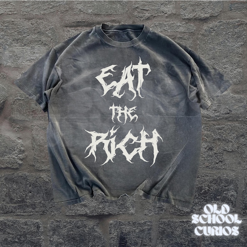 May include: A gray t-shirt with the text "EAT THE RICH" in a white, gothic font. The shirt has a vintage, washed-out look. The words are stacked vertically. The bottom right corner of the shirt has the text "OLD SCHOOL CURIOS".