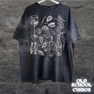 May include: Black t-shirt with a white illustration of wine, oysters, mussels, sardines, and grapes. The shirt also features two wine bottles, a corkscrew, and the text "Old School Curios" at the bottom.