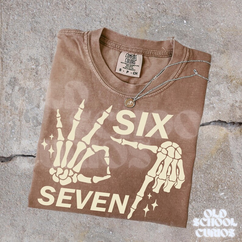 May include: A brown Comfort Colors t-shirt with a skeleton hand graphic that spells out "SIX SEVEN" in cream-colored letters. A silver necklace with a small pendant rests on the shirt. The shirt has a small tag at the neckline.